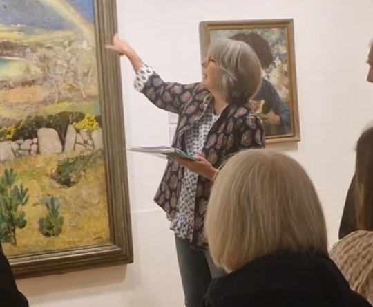 A group of people in a gallery listens to a guide describing a colorful landscape painting with a prominent rainbow. The atmosphere is attentive and educational.