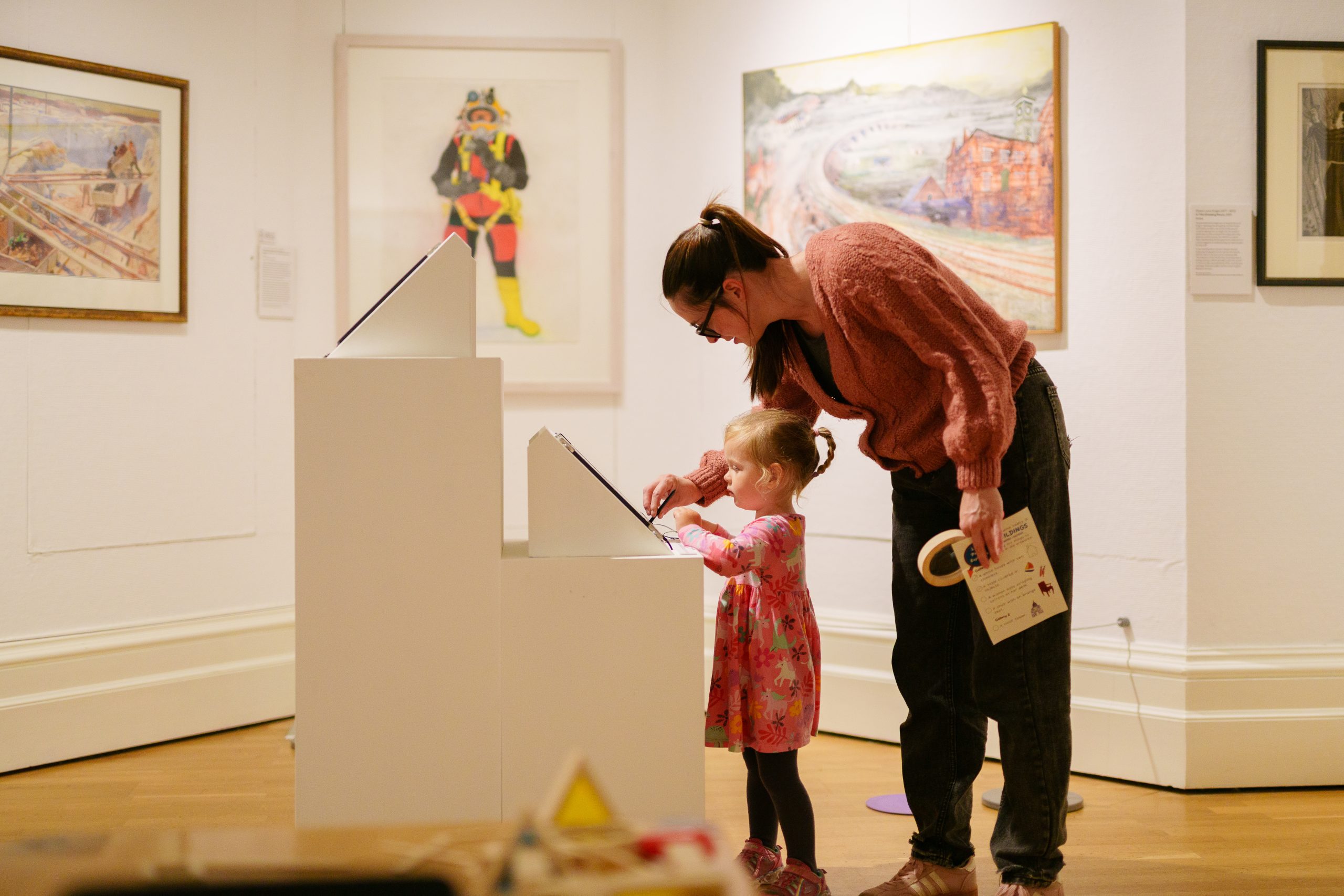 A woman and child interact with a touchscreen display in an art gallery. The child, in a pink dress, is focused, while the woman assists. Paintings hang on white walls, creating a warm, educational atmosphere.