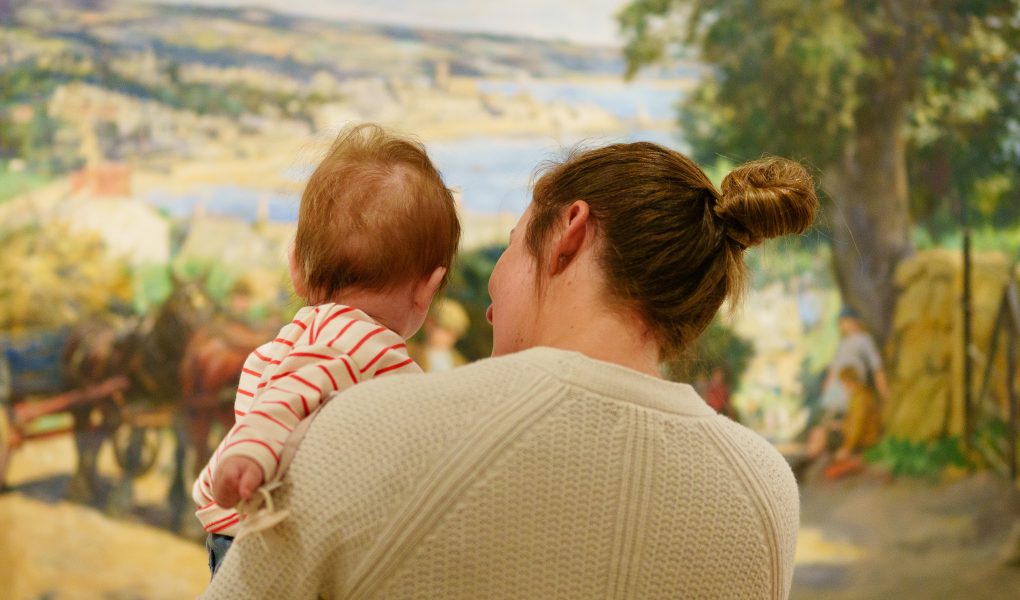 A woman holding a baby, both viewed from behind, stands in front of a colourful, pastoral painting. The scene conveys warmth and curiosity.