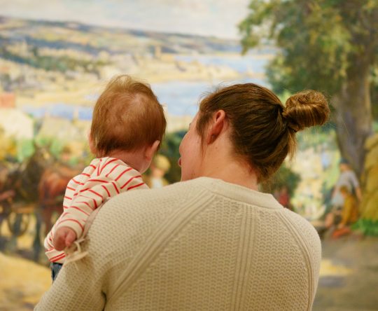 A woman holding a baby, both viewed from behind, stands in front of a colourful, pastoral painting. The scene conveys warmth and curiosity.