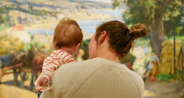 A woman holding a baby, both viewed from behind, stands in front of a colourful, pastoral painting. The scene conveys warmth and curiosity.