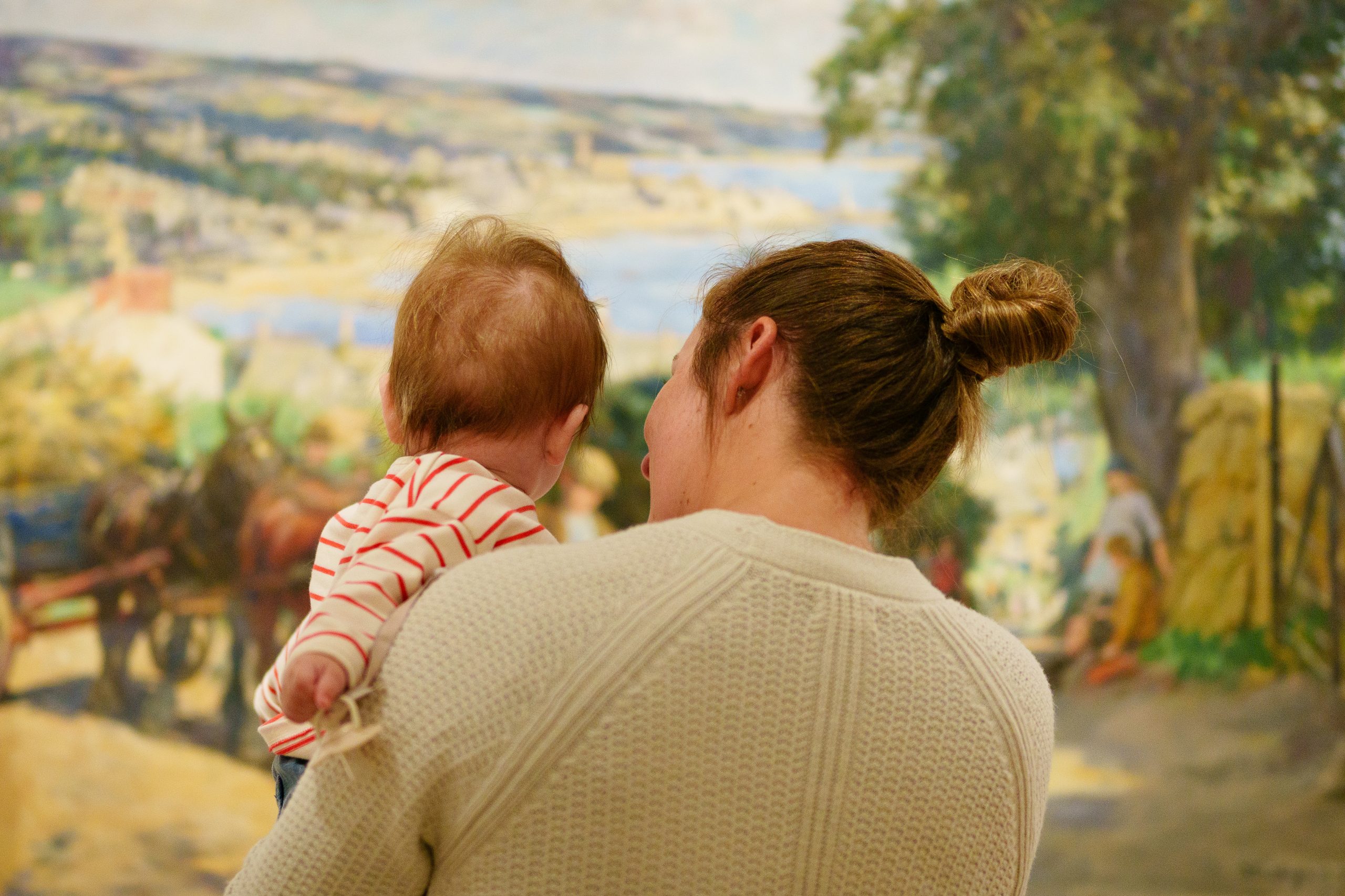 A woman holding a baby, both viewed from behind, stands in front of a colourful, pastoral painting. The scene conveys warmth and curiosity.