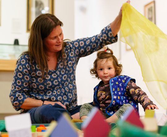 A woman and a young child play on the floor with colourful scarves and toys. The woman smiles at the child, creating a joyful and playful atmosphere.
