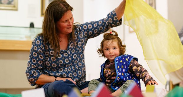 A woman and a young child play on the floor with colourful scarves and toys. The woman smiles at the child, creating a joyful and playful atmosphere.