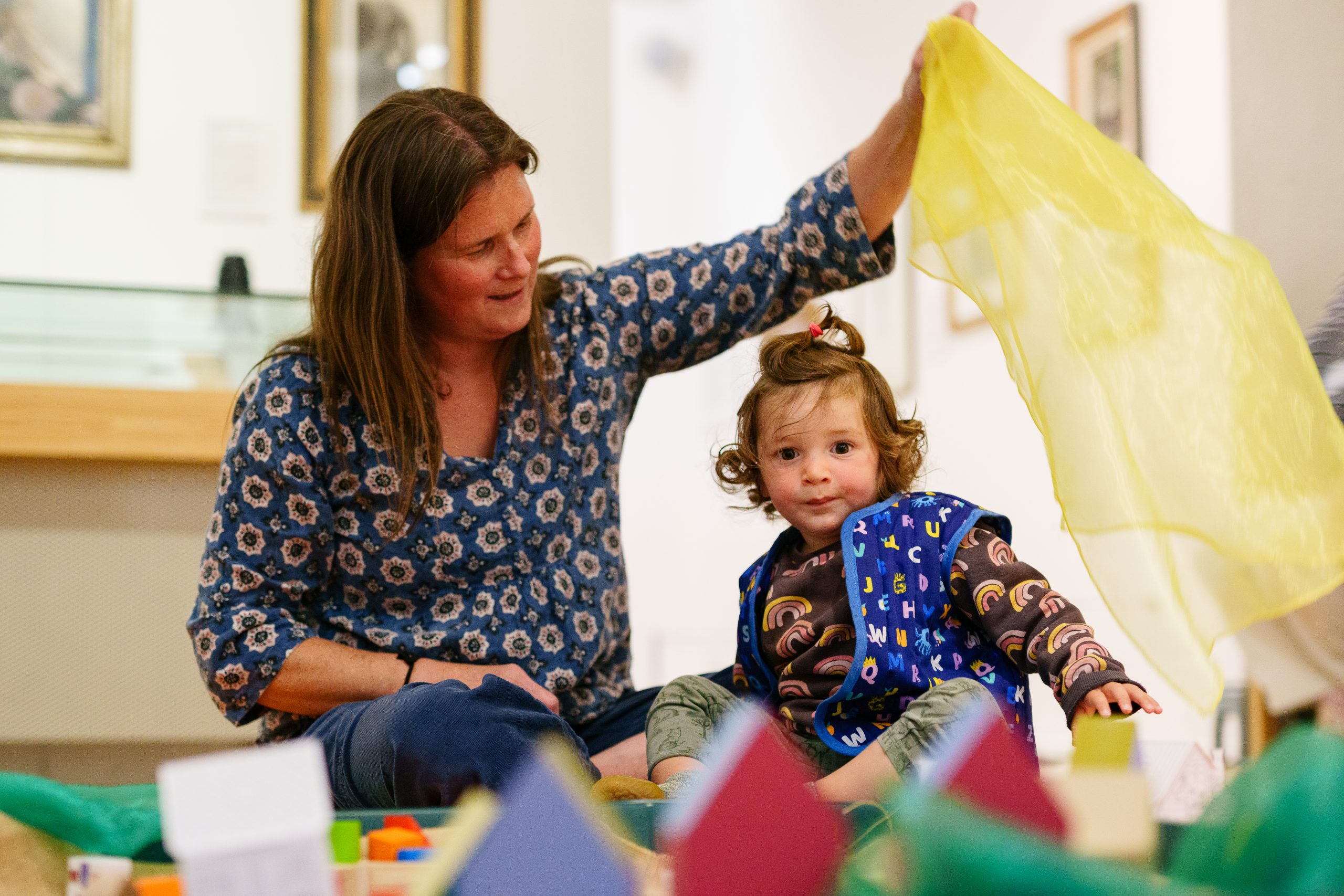 A woman and a young child play on the floor with colourful scarves and toys. The woman smiles at the child, creating a joyful and playful atmosphere.
