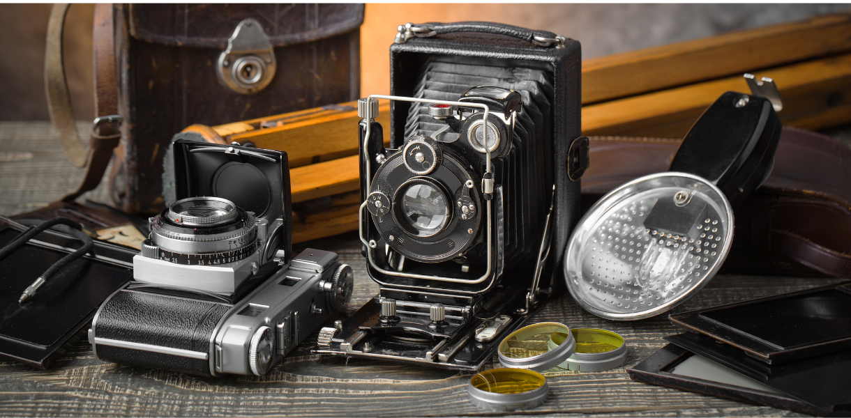 Vintage cameras and equipment on a wooden table, including a folding camera, twin-lens reflex camera, lens filters, and a leather bag, conveying nostalgia.