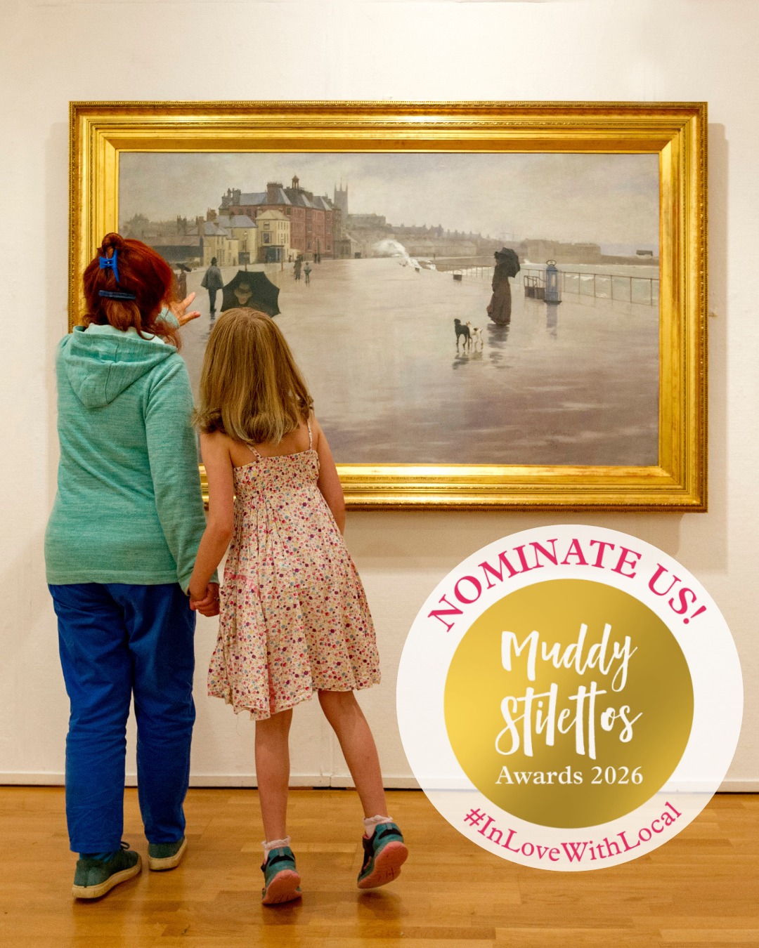 A lady and child viewed from behind stand in an art gallery looking at Norman Garstin’s 1889 painting The Rain It Raineth Every Day, a misty seaside promenade scene with figures, umbrellas, and wet pavement; promotional badge reads “Nominate Us! Muddy Stilettos Awards 2026 #InLoveWithLocal.