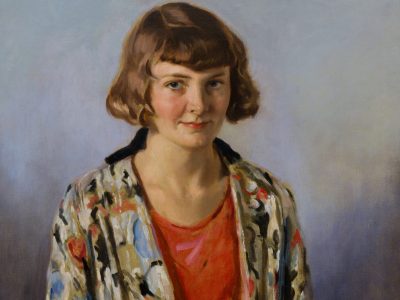 A woman with short brown hair, wearing a vibrant patterned jacket over a red dress, sits calmly against a neutral background, exuding a serene and thoughtful demeanour.