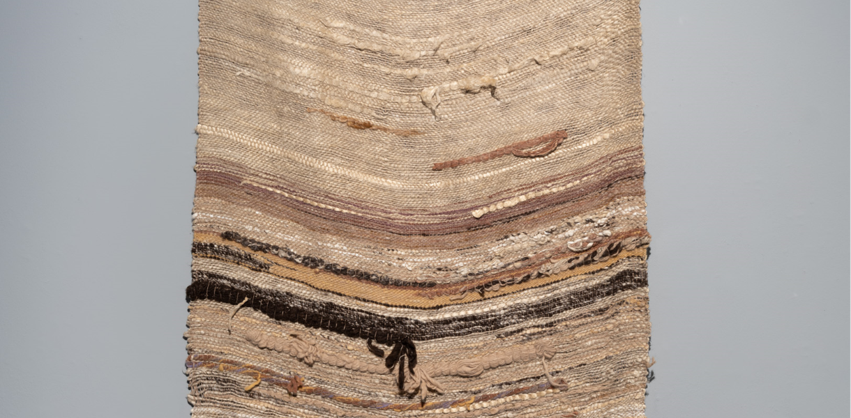 A woven textile wall hanging features earthy tones of beige, brown, and grey in horizontal layers, with textured patterns evoking a serene, natural feel.