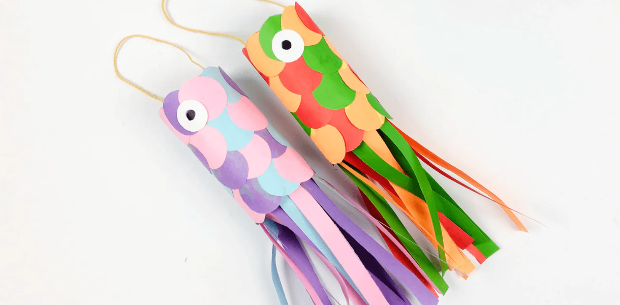 Two colourful fish crafts made from paper tubes hang by strings. Each has layered paper scales and flowing tail ribbons in vibrant hues, creating a playful and cheerful look.