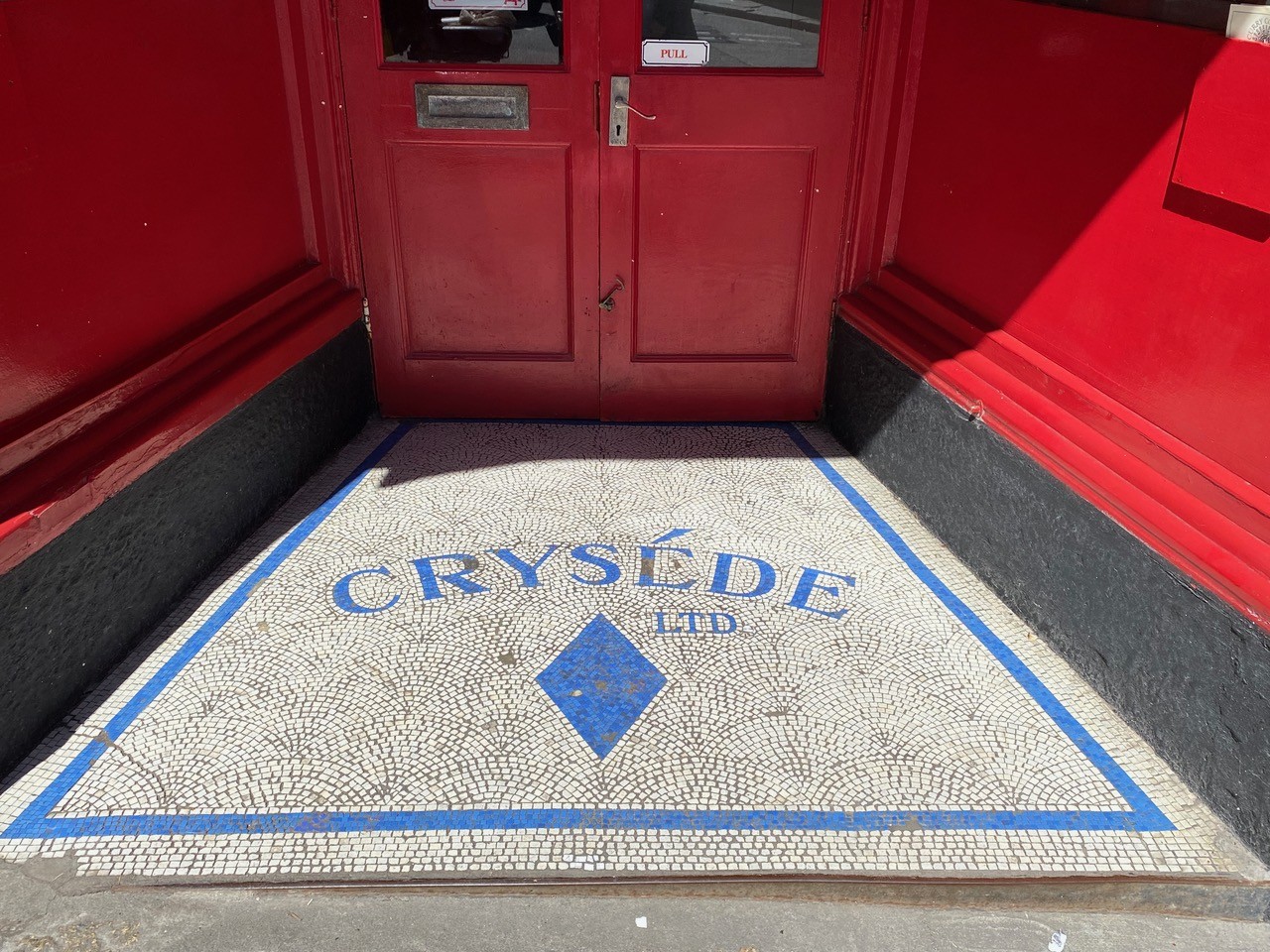 Red double doors with a mosaic tile entryway reading "CRYSÉDE LTD" in blue. The scene is vibrant, with a welcoming and artistic tone.