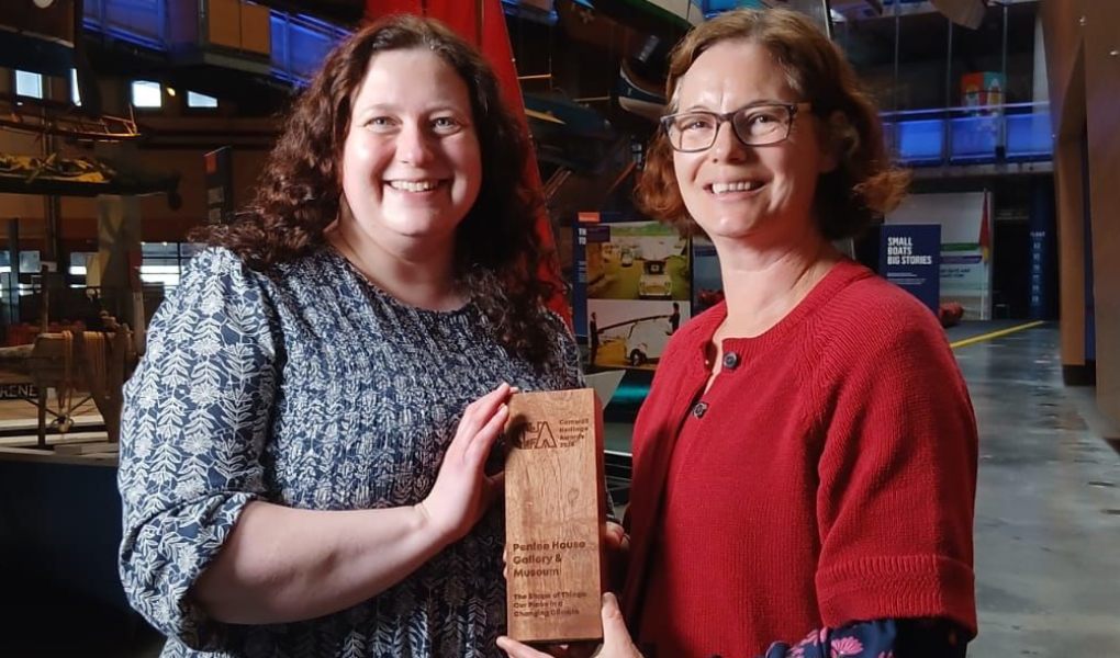 Two smiling women stand indoors holding a wooden award. Behind them are boats suspended from the ceiling. The atmosphere is celebratory.