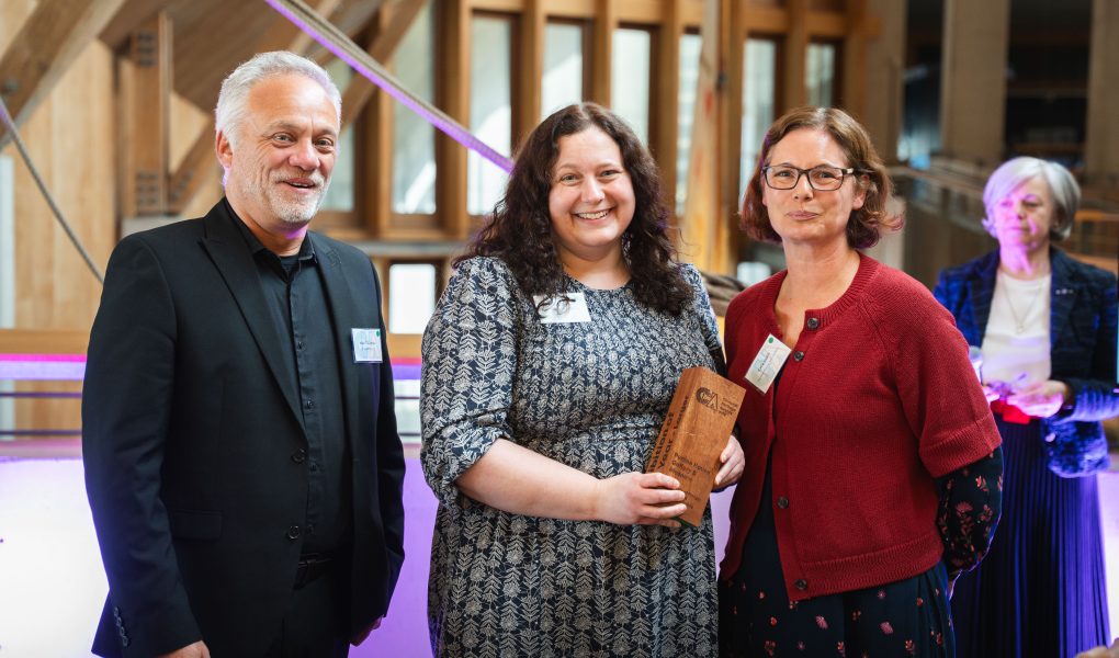 Three people smile indoors, one holding an award. Warm lighting and architectural details in the background create a celebratory atmosphere.