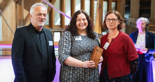 Three people smile indoors, one holding an award. Warm lighting and architectural details in the background create a celebratory atmosphere.