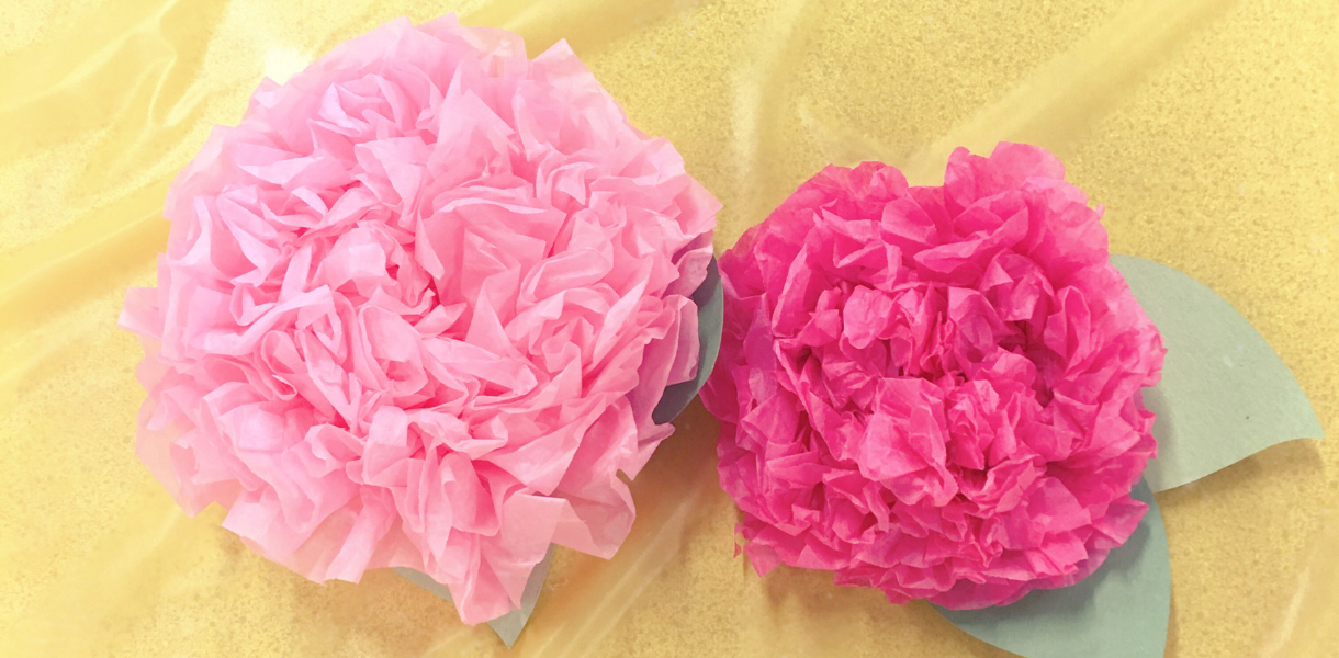 Two handcrafted tissue paper flowers on a light fabric background. The left bloom is light pink, and the right is bright pink, each with green paper leaves.