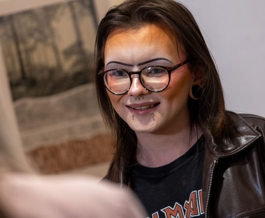 A person with glasses and facial piercings smiles warmly. They wear a leather jacket and a graphic tee. Soft focus art hangs in the background, creating a casual and friendly atmosphere.