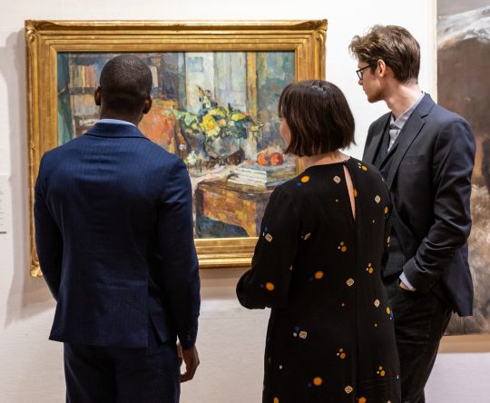 Three people view an impressionist painting in a museum. The scene is thoughtful, with a colourful artwork in a gold frame on a white wall.