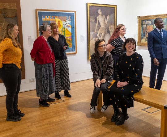 A group of people in an art gallery, smiling and engaged. They stand and sit on a bench, surrounded by colourful paintings, creating a lively and joyful atmosphere.
