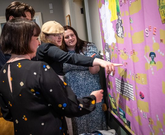 A group of people enthusiastically discusses a vibrant, collage-style art piece on a wall. They point at various elements on the colourful display.