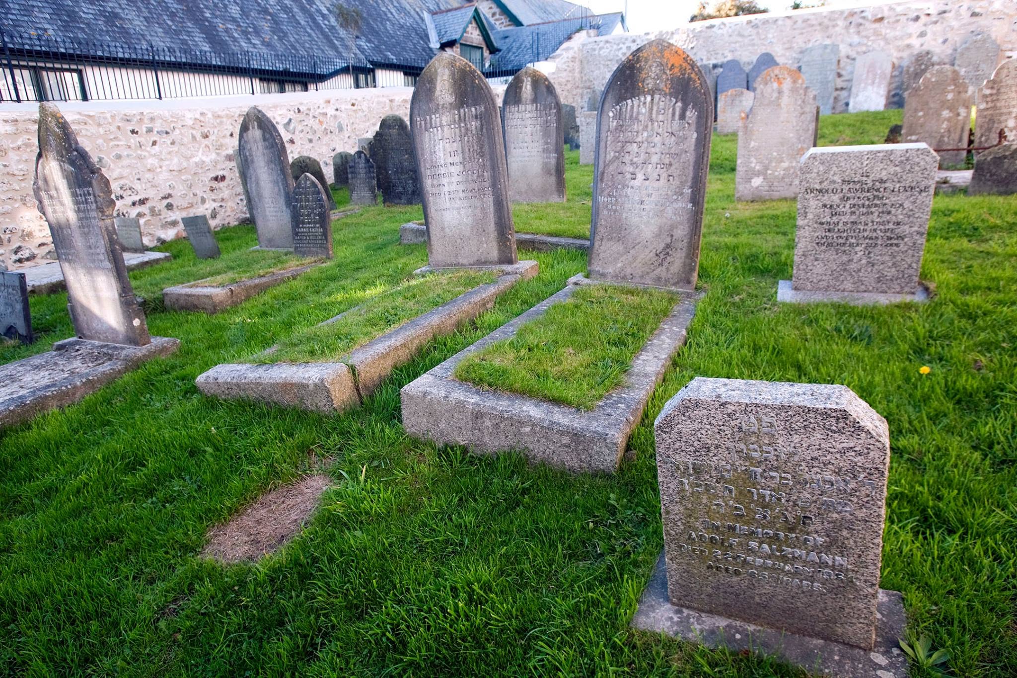 A historic Jewish cemetery with weathered headstones, inscribed in Hebrew, set amidst neatly trimmed grass and enclosed by a stone wall. Peaceful ambiance.