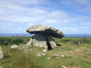 Ancient stone dolmen under a bright blue sky, surrounded by green grass and wildflowers, with the sea visible in the distant background.