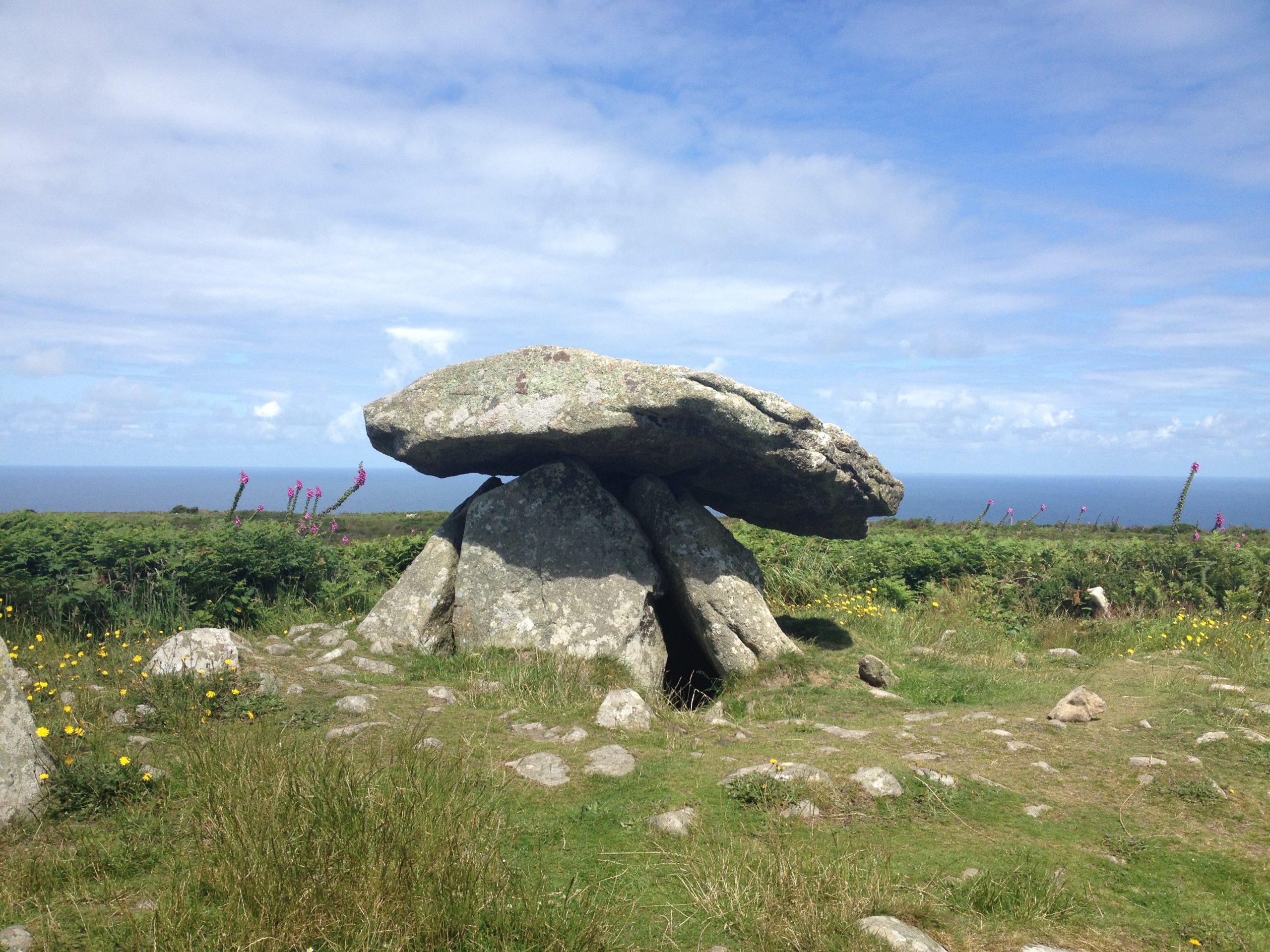 Ancient stone dolmen under a bright blue sky, surrounded by green grass and wildflowers, with the sea visible in the distant background.