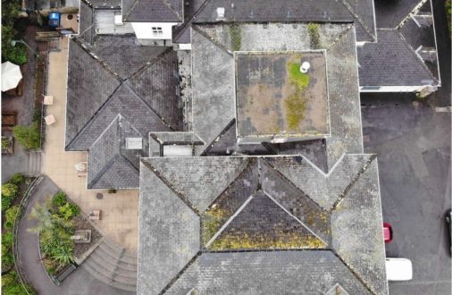 Aerial top-down view of a large building with a complex gray slate hip roof, showing weathered textures, moss, and surrounding paved patio areas.
