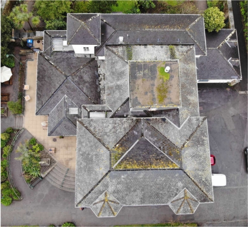 Aerial top-down view of a large building with a complex gray slate hip roof, showing weathered textures, moss, and surrounding paved patio areas.