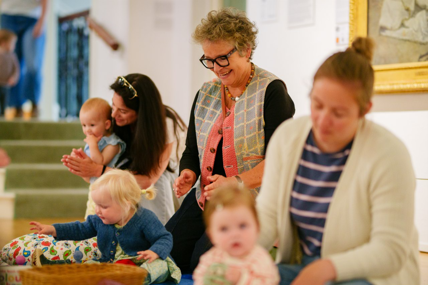 A joyful gathering in a bright room, featuring adults and babies engaged in an activity. The mood is playful, with people smiling and interacting.