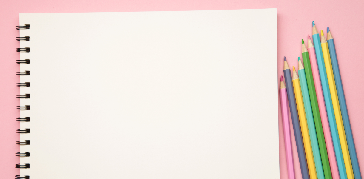 A spiral-bound sketchbook with blank white pages sits on a pink surface next to a row of colorful sharpened pencils arranged in a diagonal line.