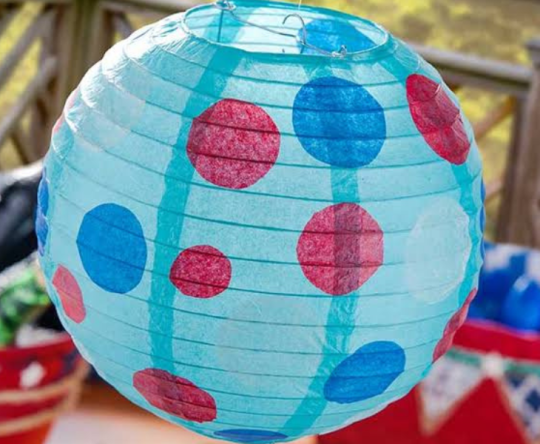 A bright turquoise paper lantern with red and blue polka dots hangs outdoors above colorful party decorations, creating a cheerful, festive mood.