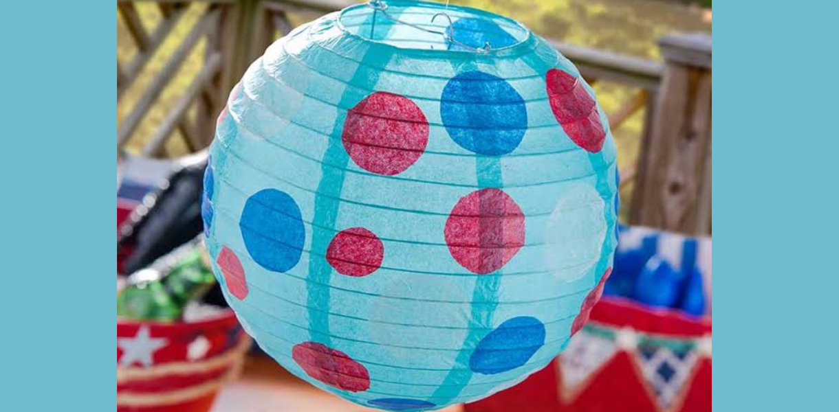 A bright turquoise paper lantern with red and blue polka dots hangs outdoors above colorful party decorations, creating a cheerful, festive mood.