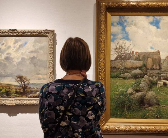 A woman with short brown hair seen from behind, standing in an art gallery and looking at two framed landscape paintings hanging on a white wall.