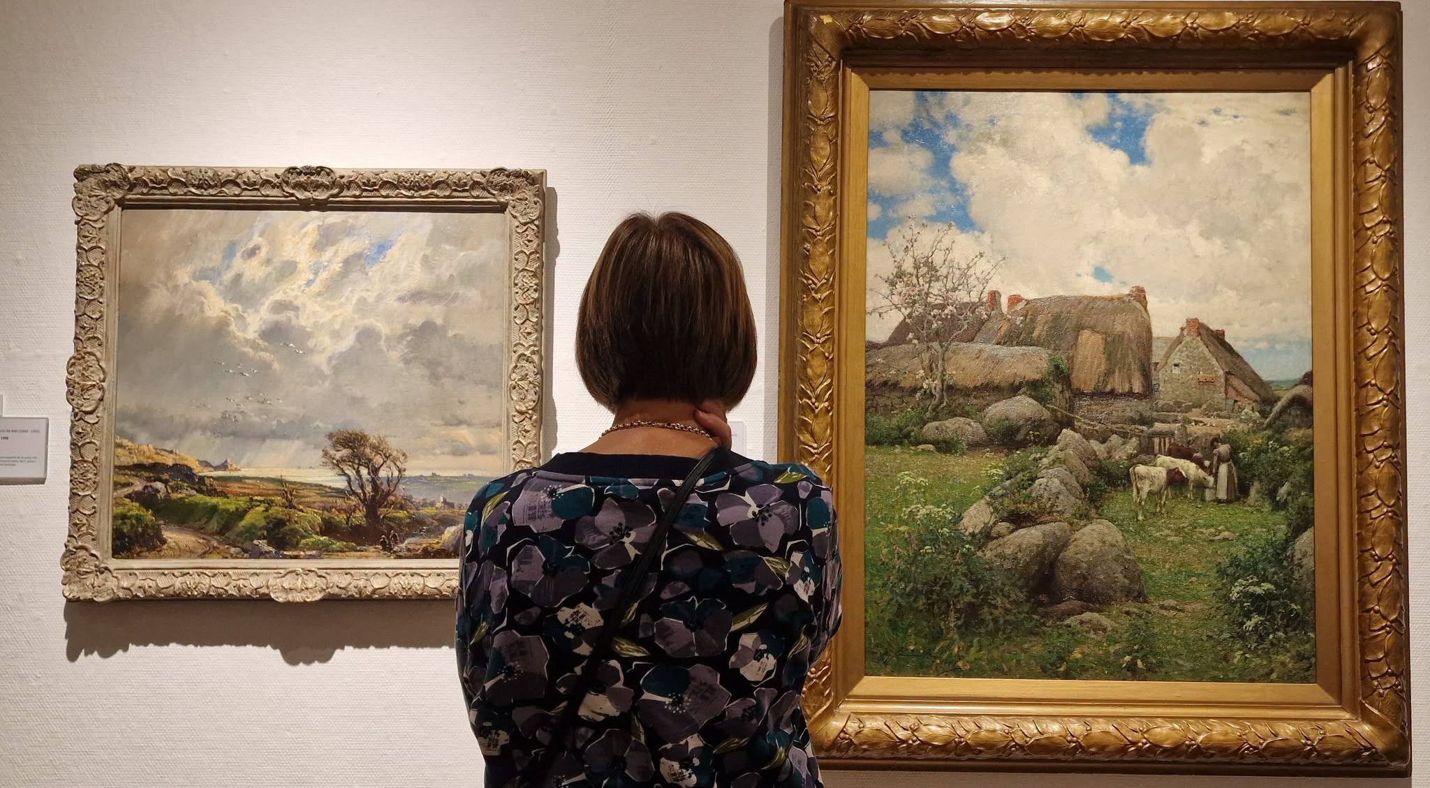 A woman with short brown hair seen from behind, standing in an art gallery and looking at two framed landscape paintings hanging on a white wall.